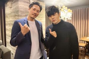 David Johnson Jiang standing indoors next to a well-known super star - Jay Chou, both facing the camera and making a hand gesture, in a warmly lit room with curtains and a chandelier in the background.