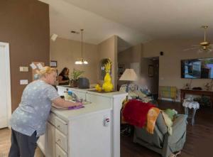 Caregiver working in the kitchen of a residential senior care home with residents relaxing in the living room.