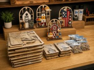 Stacks of laser-cut wooden craft kits and small bags of puzzle pieces on a workshop table next to finished, hand-painted wooden table decorations featuring a birdhouse, lighthouse, barn, and patriotic theme
