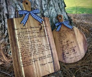 Two custom engraved wooden cutting boards with handwritten family recipes etched into the wood, tied with blue and white plaid ribbons, resting against a tree trunk outdoors
