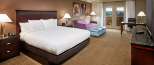 Hotel room with king bed, purple sofa, ottoman, desk, lamps, and balcony overlooking greenery and mountains.
