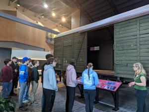 students interact with a tour guide at the Zekelman Holocaust Memorial Center, who is explaining the cable car in front of them