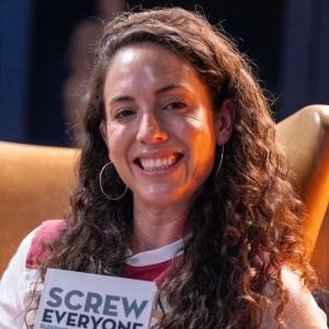 Caitlin Peluffo poses in a high-backed leather chair, holding a copy of "Screw Everyone, Sleeping My Way to Monogamy" by Ophira Eisenberg.