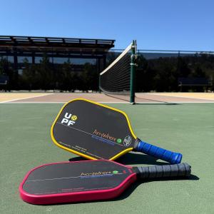 Two black Freedom Fighter Paddles by Anywhere Pickleball on a green outdoor court, one with a yellow UPF edge guard and blue grip, the other with a red edge guard and gray grip, with a pickleball net and sunny sky in the background.