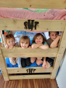 This National Make-A-Will Month, Sleep in Heavenly Peace (SHP), the leading national nonprofit dedicated to building and delivering beds to children in need, is encouraging people to consider how planned giving can create a legacy of hope Four young children smiling and peeking over the rails of a wooden bunk bed marked with the SHP logo, with pink and blue bedding visible.