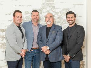 Four male real estate agents dressed in business casual attire, smiling and standing together in front of a distressed white brick wall.