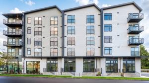 Front elevation of the Cartwright Apartments in daylight, a five-story white building with three gabled roof sections, black-framed windows, balconies and ground-floor units with individual stair entries, under a blue sky.
