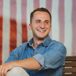 Aaron Cherry, Roseburg real estate broker, seated outdoors in a denim shirt and smiling, with an American flag mural behind him.