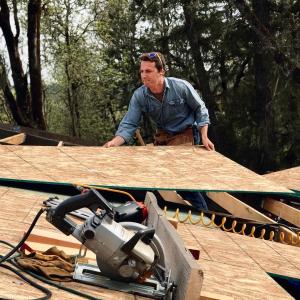 Aaron Cherry in a denim work shirt and tool belt, wearing ear protection and sunglasses pushed up on his head, standing at the edge of a roof deck sheathed in oriented strand board. A circular saw, nail gun, air hose and work gloves rest on the sheathing
