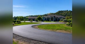Curving freshly paved black asphalt road climbing through a Northern California hillside, with an asphalt paver visible at the left edge and a yellow vibratory roller in the middle distance operated by a worker in an orange hi-vis vest, framed by oak tree