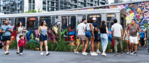 People walking past a colorful graffiti-covered train at a city station with modern buildings in the background.
