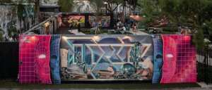 Vibrant street art mural with geometric patterns, abstract human figures, and neon grid faces on an outdoor wall in an urban park setting.