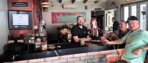 Smiling bartender behind counter serving drinks to two men and one woman seated, with framed art and decor in a cozy bar interior.