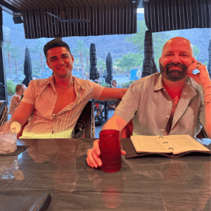 Two men smiling and sitting at an outdoor restaurant table with mountains and umbrellas in the background.