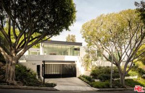 A modern, multi-level house with sleek white walls and extensive floor-to-ceiling glass paneling is viewed from the street. A secure black gate frames the entrance to the concrete driveway, all bordered by two large, overarching trees.