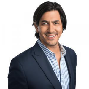 A professional portrait of a smiling male real estate agent with medium-length dark hair, wearing a navy blue blazer over a light blue and white striped shirt.