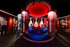 Visitors seated in the red-lit Great Depression gallery at the redesigned Herbert Hoover Presidential Library and Museum, surrounded by Market Crash, Banking Crisis, and Farming Crisis exhibit panels facing a theatrical stage set.