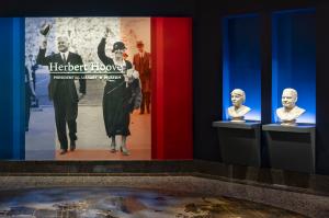 Matching marble busts of Herbert and Lou Henry Hoover displayed side by side at the entrance to the redesigned Herbert Hoover Presidential Library and Museum, beneath a large photograph of the couple waving to a crowd.
