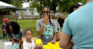A young girl collects supplies at a distribution table while a volunteer in a 1st Choice Family Services shirt helps hand out items.