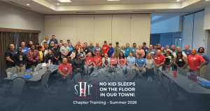 Group photo of dozens of Sleep in Heavenly Peace chapter volunteers and leaders standing and seated around conference tables in a training room, smiling for a group photo. Text overlay reads 