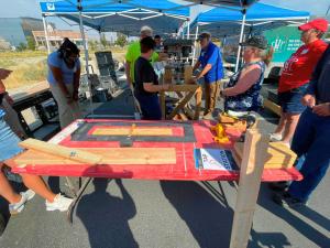 Volunteers gather around an outdoor workstation under a blue canopy tent, assembling wooden bed frame pieces on a red table labeled 
