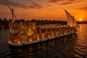Kenzy Dahabeya, a luxury boutique Nile cruise boat, sailing at sunset on the Nile River with traditional sails, warm golden lighting, and lush palm trees lining the riverbank between Luxor and Aswan.