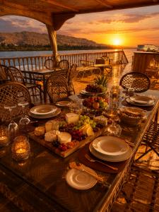 Elegant open-air dining deck aboard Kenzy Dahabeya featuring a gourmet dinner table with artisan cheeses, fresh fruit, candles, and fine tableware overlooking the Nile River at sunset, with panoramic views of the river and surrounding mountains.