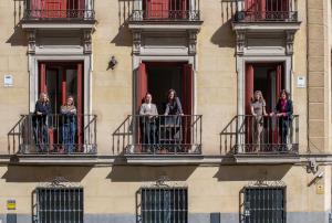The team of Bacana Communications stands on three adjoining wrought-iron balconies of a classic Madrid building, with red-shuttered windows and a beige stone façade, on a sunny day in Spain.  Title tag: Bacana Communications — Madrid, Spain Agency Office