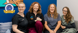 Four smiling women in medical scrubs sit on a couch under a Certified Autism Center badge on a blue and gray wall background.