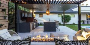 Phoenix AZ property owners are booking outdoor projects earlier than usual this year Covered outdoor kitchen with a stone island, built-in grill and pizza oven beside a lit fire pit and lounge seating at a Phoenix Arizona home at dusk