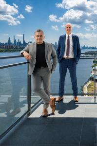 Enrique Napoles and Mark Trompeter standing on the terrace of the 1000 Portside Drive penthouse in Edgewater, New Jersey, with the Hudson River and Manhattan skyline behind them.