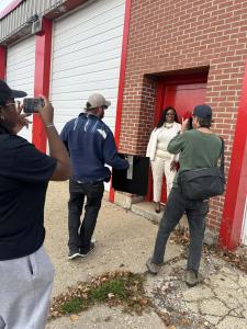 Natasha Broxton standing outside the Select Auto Parts facility while a media team photographs her.