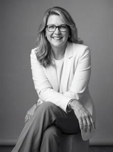Author Melissa Fisher wears a cream jacket and tan pants sits on stool arms folded leaning forward smiling after completing her manuscript SIGNAL