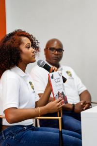 Kylar Denae Williams of Legacy Health Works speaking into a microphone while holding her book Two Homes, One Heart, with Kevin D. Williams seated behind her.