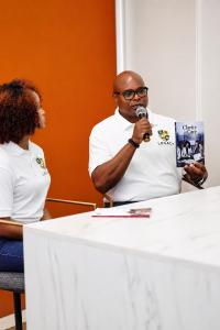 Kevin D. Williams of Legacy Health Works speaking into a microphone while holding his book Clarity in Care, with Kylar Denae Williams seated beside him.