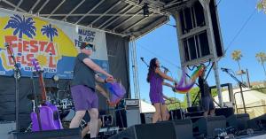Kathryn the Grape performing live at the Fiesta del Sol Music Festival in Solana Beach, California, on May 30, 2026. Wearing purple, she sings on an outdoor stage while leading an interactive performance with colorful rainbow scarves, accompanied by perfo
