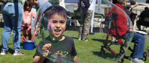 A young boy wearing headphones smiles inside a large soap bubble at an outdoor event with people and chairs in the background.