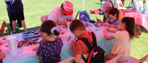 Children and adults seated at a table outside, engaged in drawing and coloring activities under a tent.
