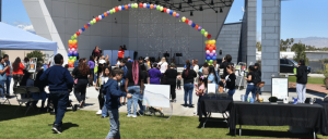 Community outdoor event with families, balloon arch, children playing with bubbles, and people gathered near a stage on a sunny day.