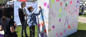 Children and teens draw colorful pictures and write messages on large white panels outdoors at a sunny event.