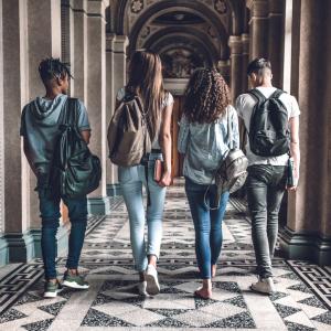 Four college students wearing backpacks walk together through an arched campus hallway.