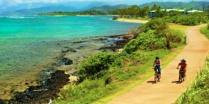 couple rides bikes on a winding bike path, with the ocean nearby and mountains in the distance