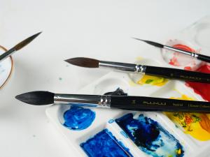 Close-up of FUUMUUI watercolor brushes resting on a palette with blue, yellow and red watercolor paint.