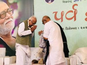 Two guests exchanging a traditional namaste greeting on stage during a cultural event, with a large portrait and Gujarati text visible in the background.