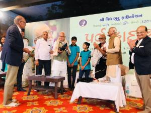 Group of distinguished guests, community members, and children standing together on stage at a Shri Vidyamandir Foundation event, with Gujarati text and a large portrait displayed in the background.