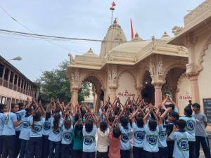 Group of students wearing matching T-shirts standing outside a Hindu temple with their hands raised in prayer, with the temple’s ornate architecture and flags visible in the background.