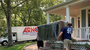 Two uniformed Arnoff employees carrying a large, wrapped piece of furniture down a walkway towards a white Arnoff Moving & Storage truck parked in a residential driveway. Image 3 (image_2.png)