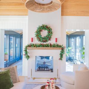Celestial At Home battery operated Woodland Christmas wreath with dual color LEDs, red berries and real pinecones hung above a white fireplace mantel with the matching pre-lit Woodland garland and red pillar candles