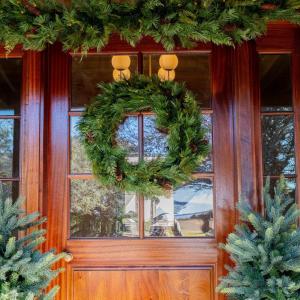 Celestial At Home battery operated Aspen Christmas wreath with dual color T5 LEDs centered on a mahogany front door beneath the matching pre-lit Aspen cedar garland, flanked by frosted mini trees