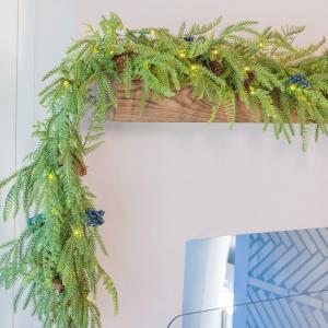 Celestial At Home battery operated Norfolk Pine Christmas garland with warm white LED fairy lights, soft fern-like tips, real pinecones and lifelike juniper blueberries draped over a wooden mantel corner